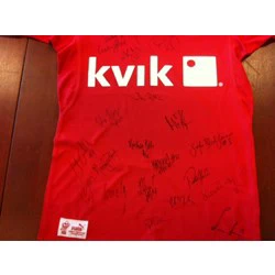 En rød skjorte med det hvide kvik-logo og flere sorte signaturer skrevet hen over fronten. Skjorten er lagt fladt på en træoverflade.